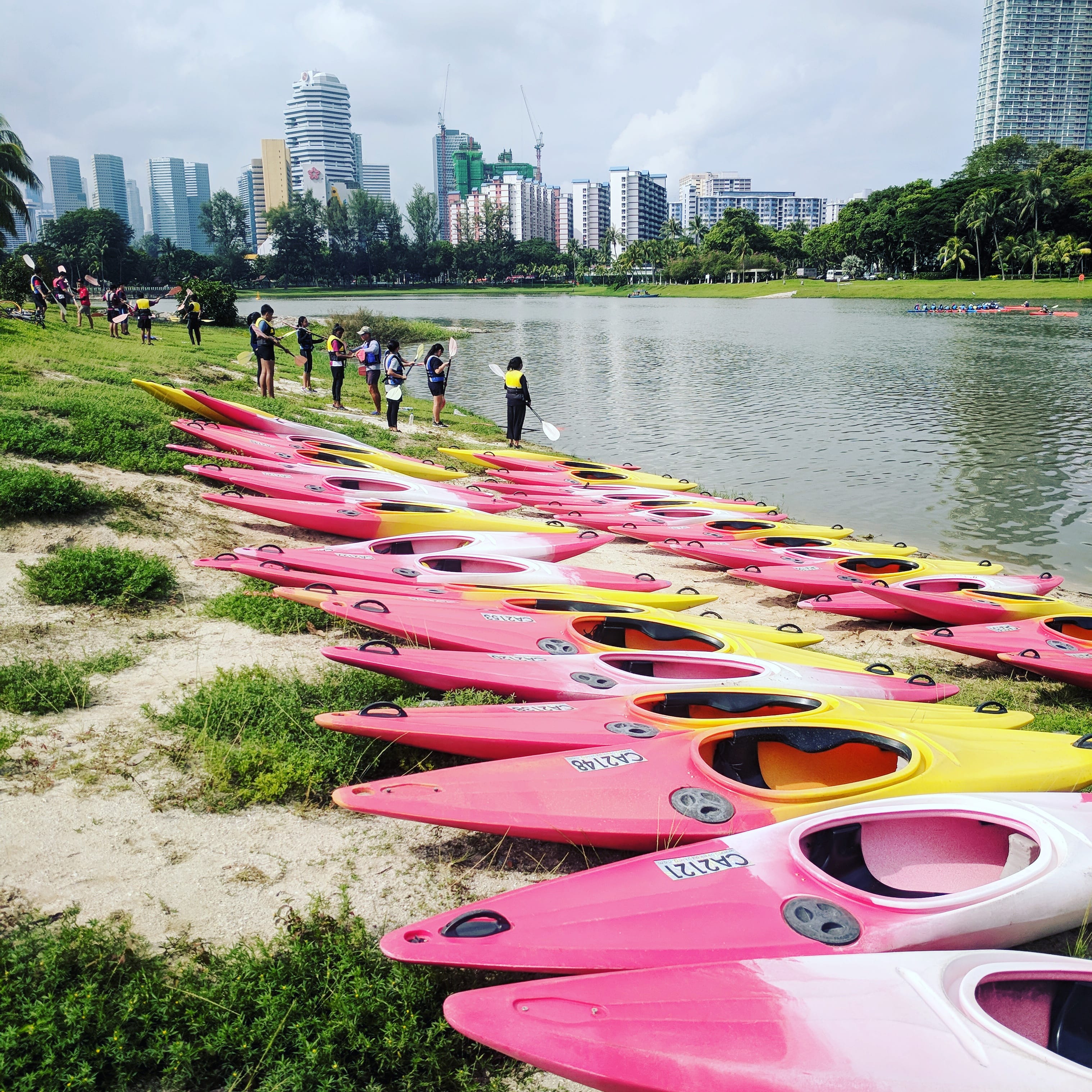 Rows of pink and yellow kayaks on a lakeshore, people in life vests, city skyline behind trees.